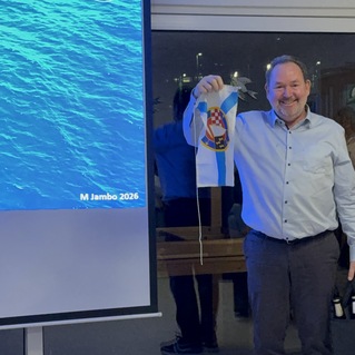 Mann h&auml;lt ein Banner mit einem Logo in einer Feier-Umgebung, im Hintergrund eine Pr&auml;sentationsfolie und applaudierende Personen.