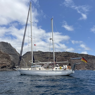 Segelboot mit deutscher Flagge schaukelt im Wasser, umgeben von felsiger K&uuml;ste unter bew&ouml;lktem Himmel.