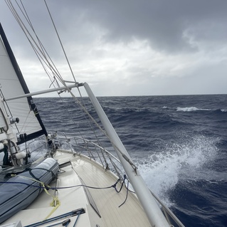 Segelboot auf offener See mit aufgew&uuml;hltem Wasser und wolkenverhangenem Himmel, Teilansicht des Bootes und Segels.