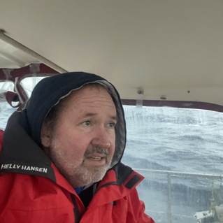 Mann in wetterfester Jacke schaut nachdenklich aus dem Fenster eines Bootes w&auml;hrend st&uuml;rmischem Wetter.