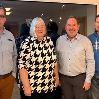 Vier Personen stehen vor einem Fenster: eine Frau mit einem schwarz-wei&szlig;en Muster und drei M&auml;nner in unterschiedlichen Oberteilen.