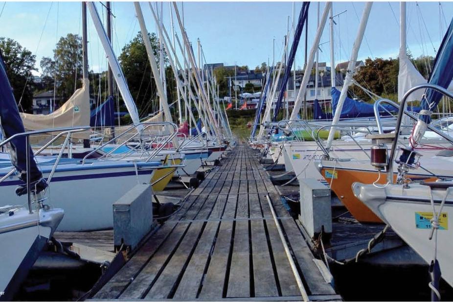Holzsteg zwischen mehreren Segelbooten im Hafen, umgeben von B&auml;umen und Geb&auml;uden im Hintergrund.