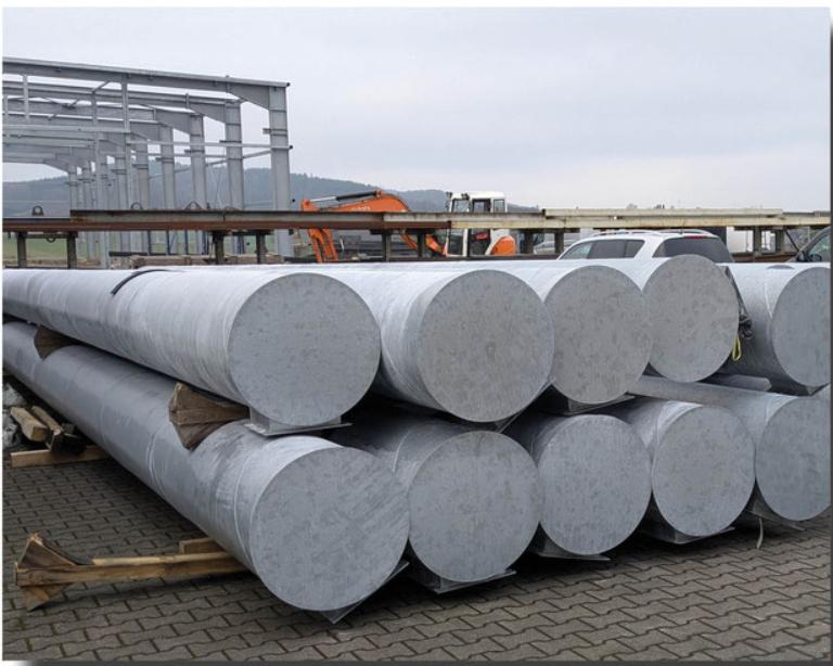 Lagerfl&auml;che mit mehreren gro&szlig;en, zylindrischen Stahlrohren auf Holzpaletten, im Hintergrund leichte Industriearchitektur.