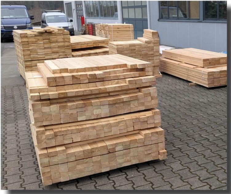 Stapeln von geklammerten Holzlatten auf einem Hof, umgeben von Lieferfahrzeugen und einem Geb&auml;ude.