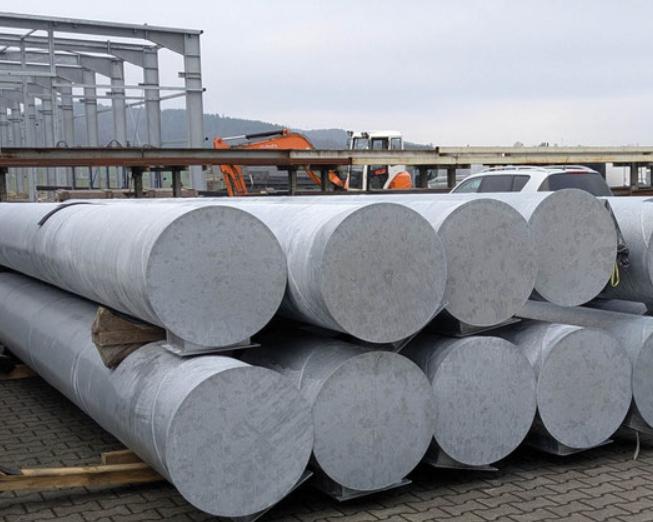 Lagerfl&auml;che mit mehreren gro&szlig;en, zylindrischen Stahlrohren auf Holzpaletten, im Hintergrund leichte Industriearchitektur.