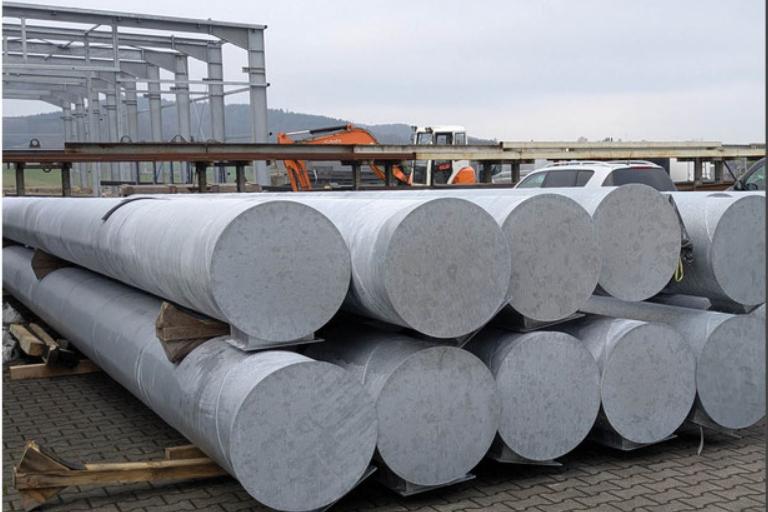 Lagerfl&auml;che mit mehreren gro&szlig;en, zylindrischen Stahlrohren auf Holzpaletten, im Hintergrund leichte Industriearchitektur.