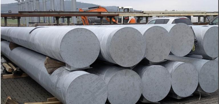 Lagerfl&auml;che mit mehreren gro&szlig;en, zylindrischen Stahlrohren auf Holzpaletten, im Hintergrund leichte Industriearchitektur.