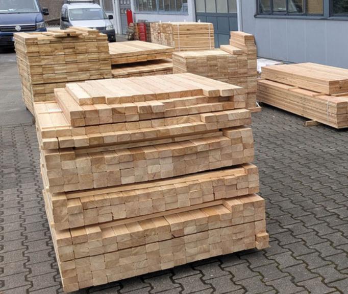 Stapeln von geklammerten Holzlatten auf einem Hof, umgeben von Lieferfahrzeugen und einem Geb&auml;ude.