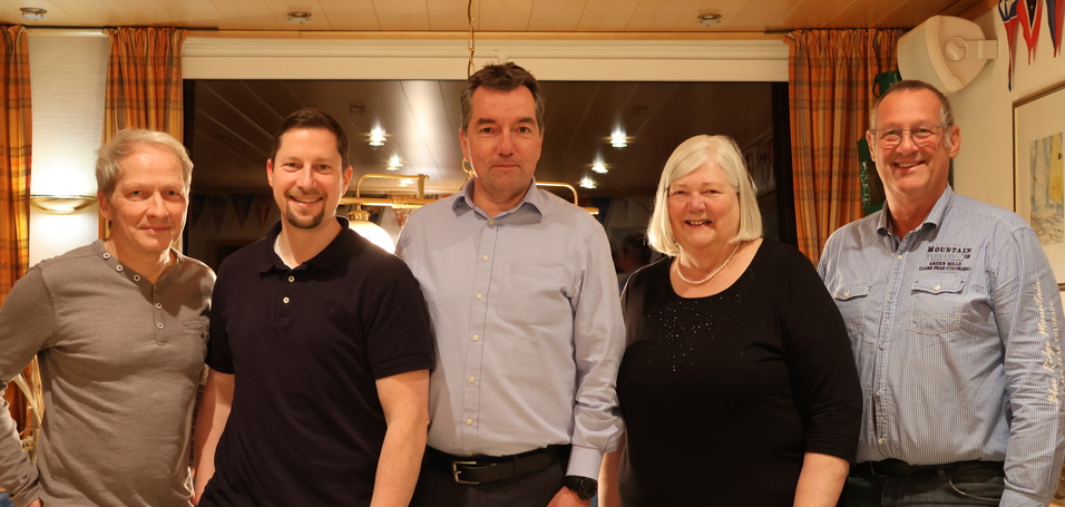 F&uuml;nf Menschen stehen l&auml;chelnd in einem Raum mit Holzverkleidung und bunten Wimpeln im Hintergrund.