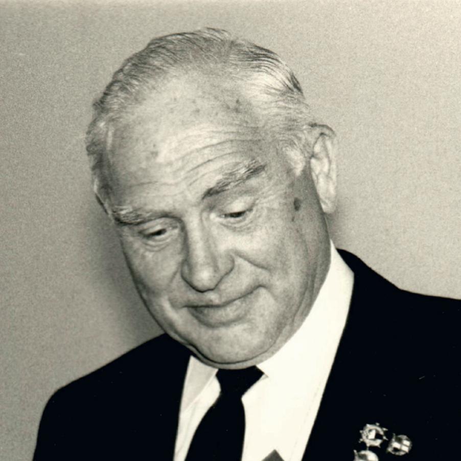 Schwarz-wei&szlig;-Foto eines Mannes in Anzug mit Abzeichen, der ernsthaft nach unten schaut.