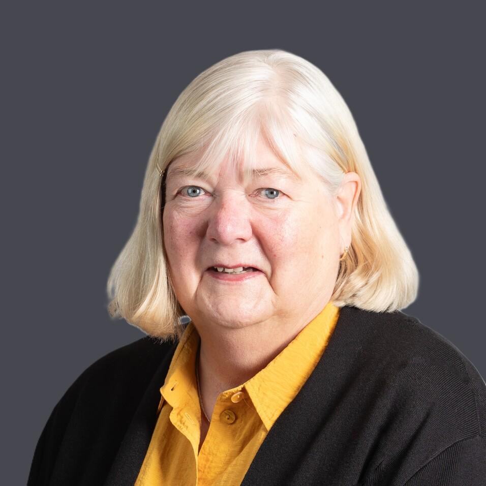 Portr&auml;t einer l&auml;chelnden &auml;lteren Frau mit blonden Haaren, die einen schwarzen Cardigan und ein gelbes Hemd tr&auml;gt.