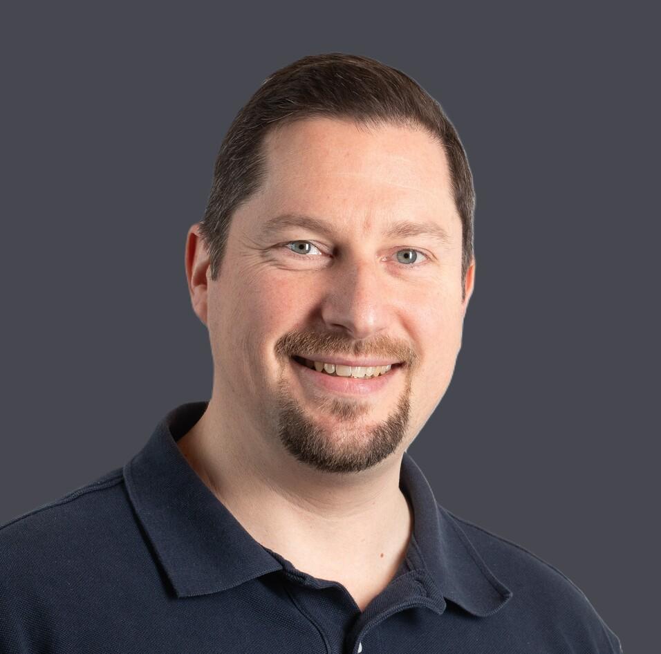 Portr&auml;t eines l&auml;chelnden Mannes mit kurzer Frisur, tragend ein dunkles Polohemd, vor einem hellen Hintergrund.