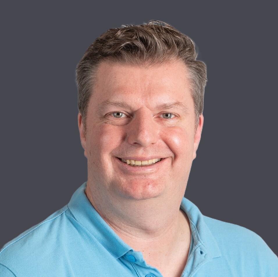 Mann mit kurzen Haaren in einem blauen Polohemd l&auml;chelt vor einem hellen, strukturierten Hintergrund.