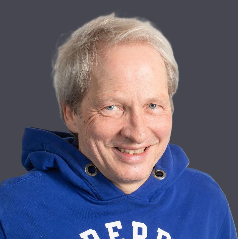 Mann mit blonden Haaren und blauer Kapuzenjacke l&auml;chelt vor einer hellen, strukturierten Hintergrundwand.