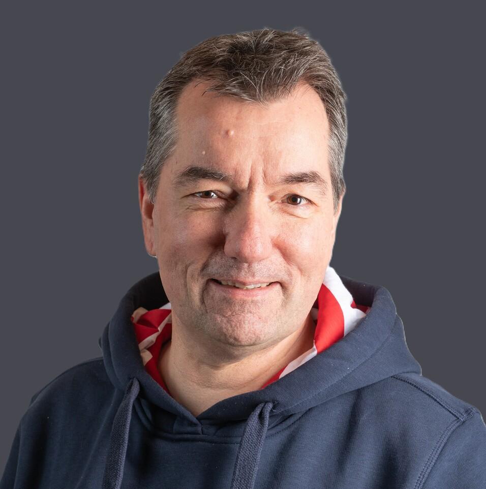 Mann mit kurzem Haar tr&auml;gt einen blauen Sweatshirt und l&auml;chelt vor einem hellen Hintergrund.