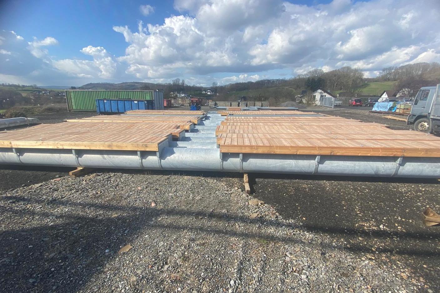 Holzplanken auf einer Metallkonstruktion, umgeben von Schotter und Himmel mit Wolken. Baustellenumgebung im Hintergrund.