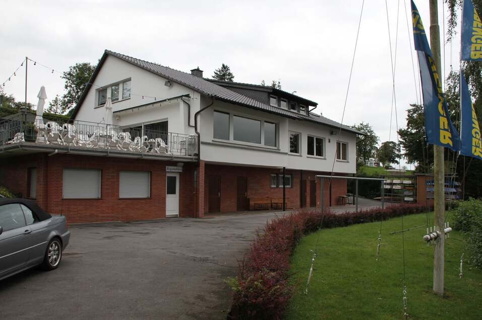 Blick auf ein modernes zweigeschossiges Haus mit Terrasse, umgeben von Grünflächen und einem gepflasterten Zufahrtsweg.