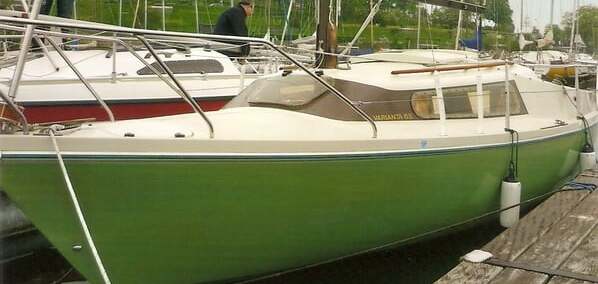 Gr&uuml;ne Segelyacht im Hafen mit zwei Personen an Bord, anderen Booten und einem Ufer im Hintergrund.