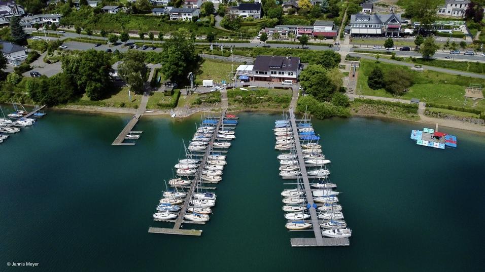 Luftaufnahme eines Yachthafens mit mehreren Booten und einem Steg, umgeben von gr&uuml;ner Landschaft und Wohngebieten.