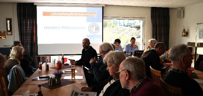 Jahreshauptversammlung mit mehreren Teilnehmern an Tischen, im Hintergrund eine Leinwand mit Informationen.