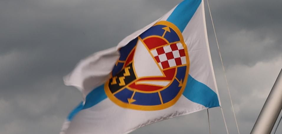 Mast mit Flaggen und Wetterfahne unter bedrohlich grauem Himmel.