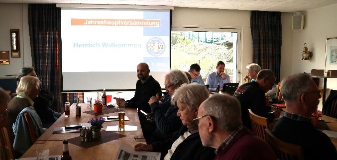 Jahreshauptversammlung mit mehreren Teilnehmern an Tischen, im Hintergrund eine Leinwand mit Informationen.