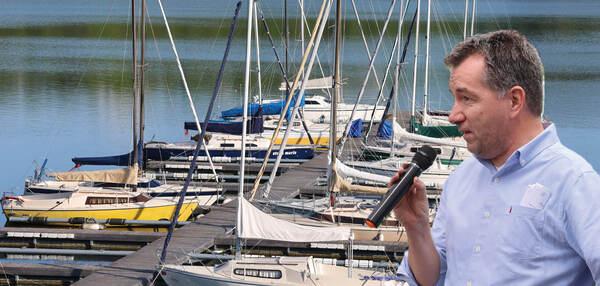 Mann spricht mit Mikrofon vor einem Hafen voller Segelboote und ruhigem Wasser im Hintergrund.