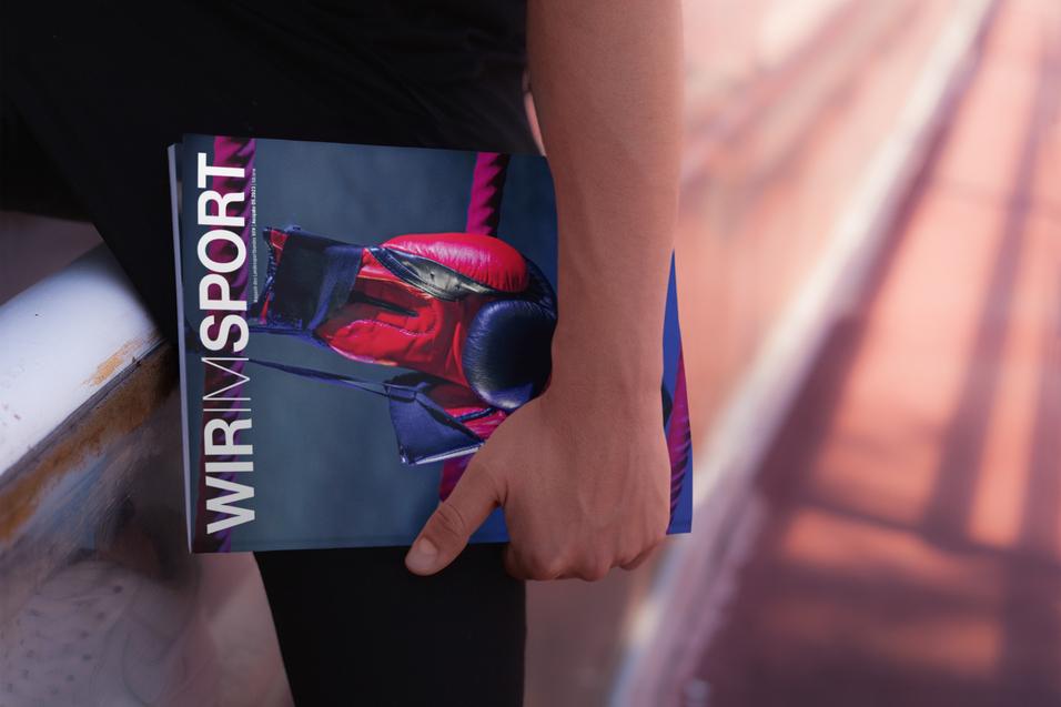Hand h&auml;lt ein Magazin mit dem Titel &bdquo;WIR IM SPORT&ldquo; auf einer Sportbahn.